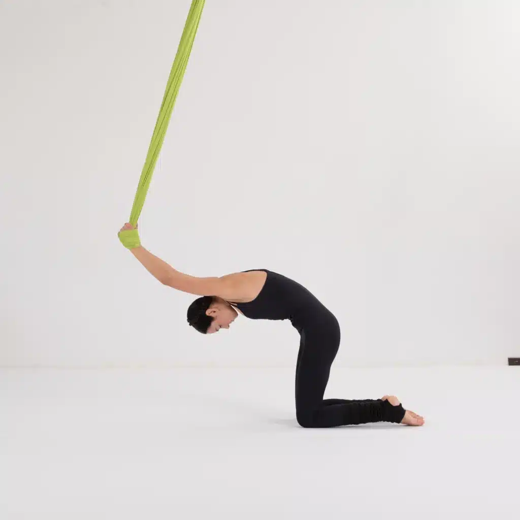 Aerial Yoga Übung im Vierfüßlerstand (Bank): Sanfte Rückendehnung mit Unterstützung des grünen Yoga-Tuchs in einem Berliner Studio.