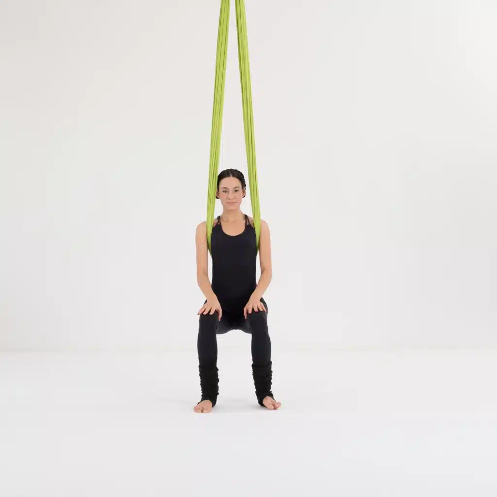 Aerial Yoga Stuhl Übung im Aerial Yoga Tuch für Zuhause. Vorderansicht der Stuhlposition mit gebeugten Knien und aufrechter Haltung zur Aktivierung von Beinen und Rücken.